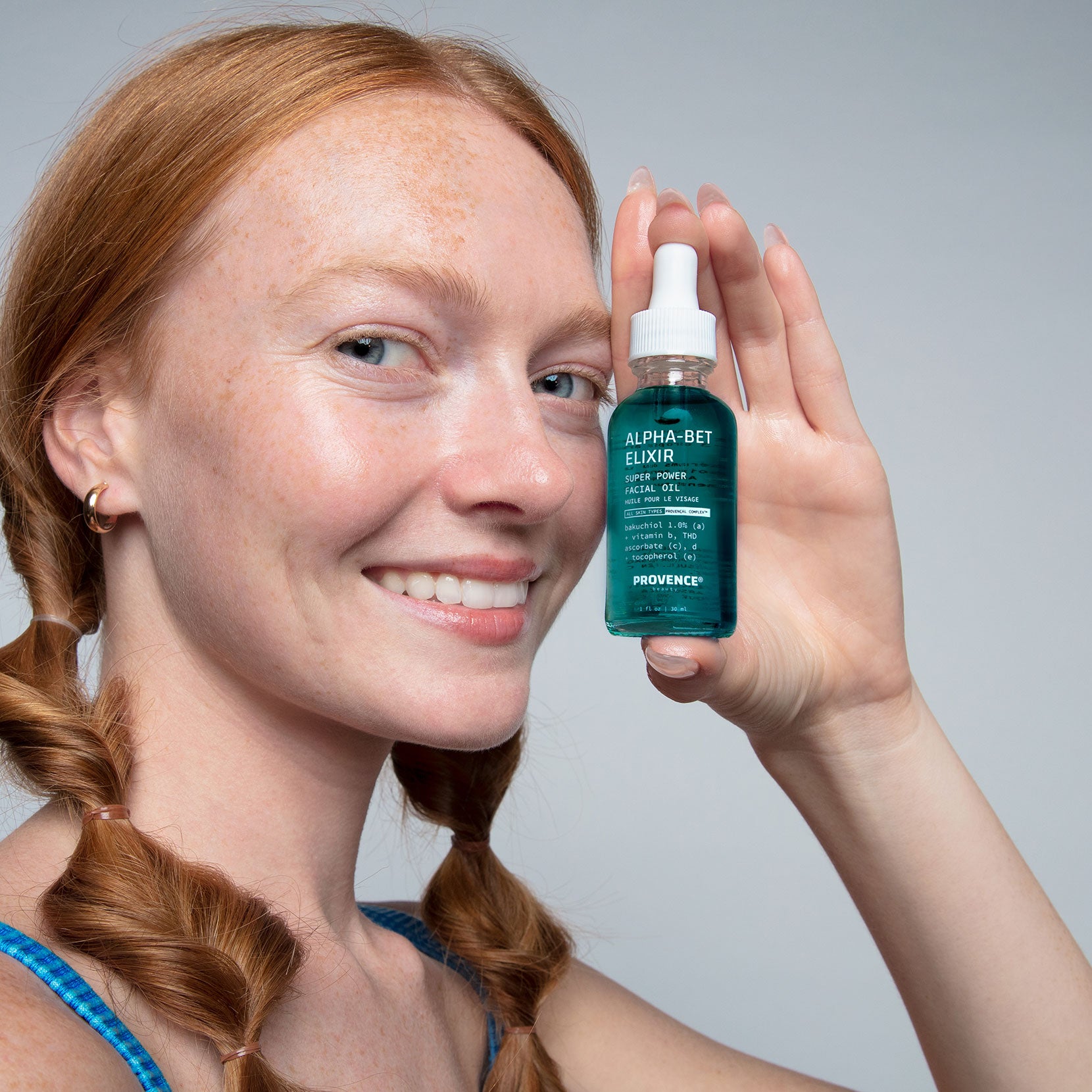 A model with glowing skin and red hair with bubble braids holding Alpha-Bet Elixir Super Power Facial Oil up by her face. Showcasing the bright turquoise gem color of the product