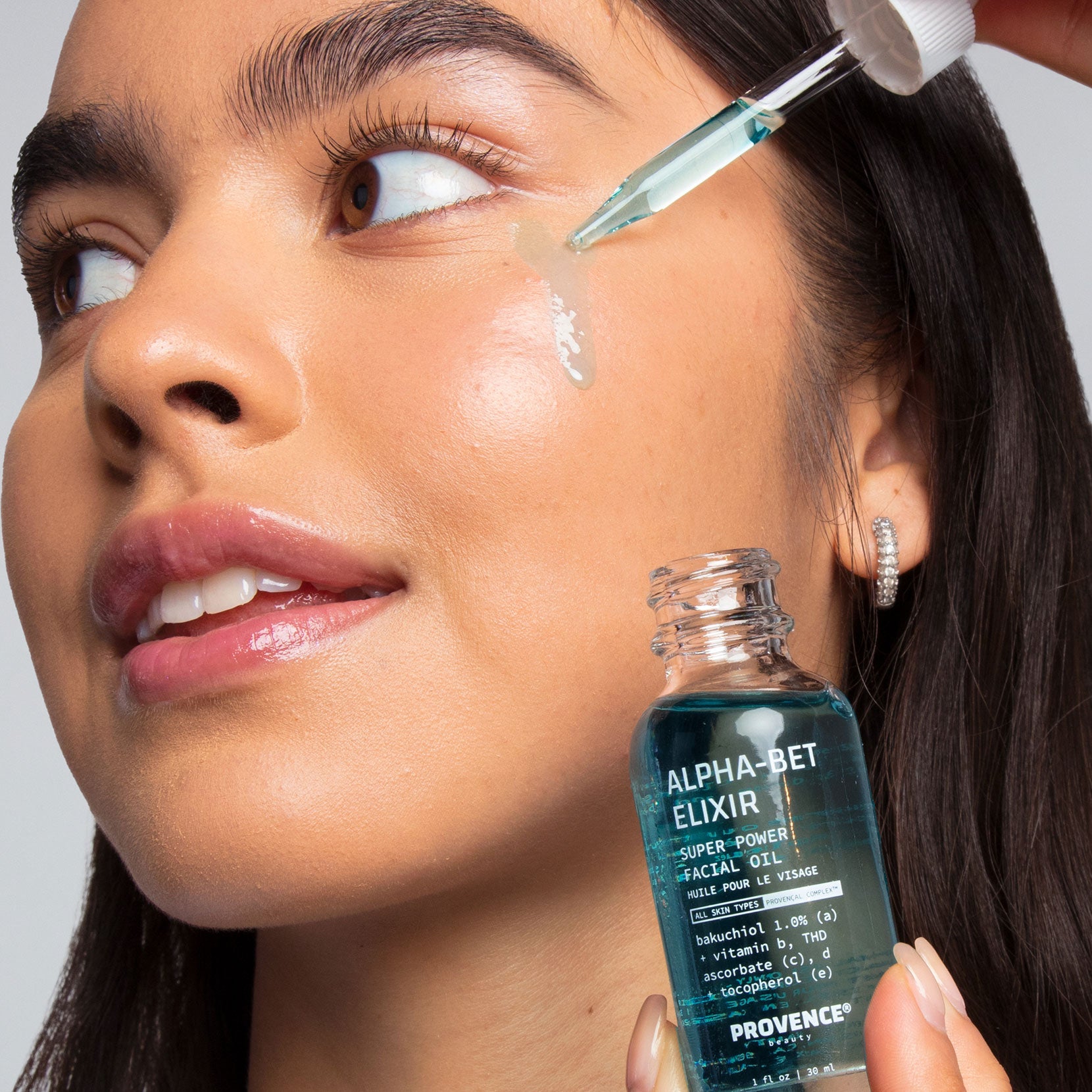 A model with glowing skin and dark brown hair applying Alpha-Bet Elixir Super Power to her cheek. The light blue color contrasts with her complexion.
