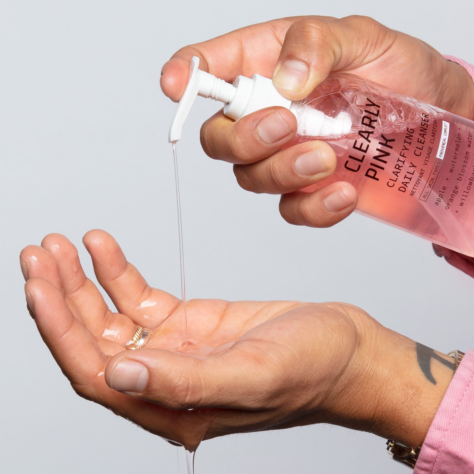 A photo of a model’s hands holding Clearly Pink Clarifying Cleanser and pressing down on the hand pump for the product liquid to run down through his hand and dripping down below. Showcasing the consistency of the product.