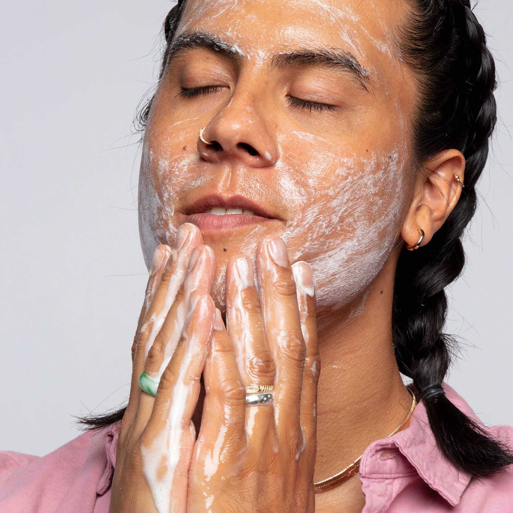 Image of a model applying the Clearly Pink Clarifying Cleanser to his face. The model’s eyes are closed and the content is around his hands, cheeks, and forehead. Due to the consistency of the cleanser there are bubbles from the products that are lathered on his face and hands.