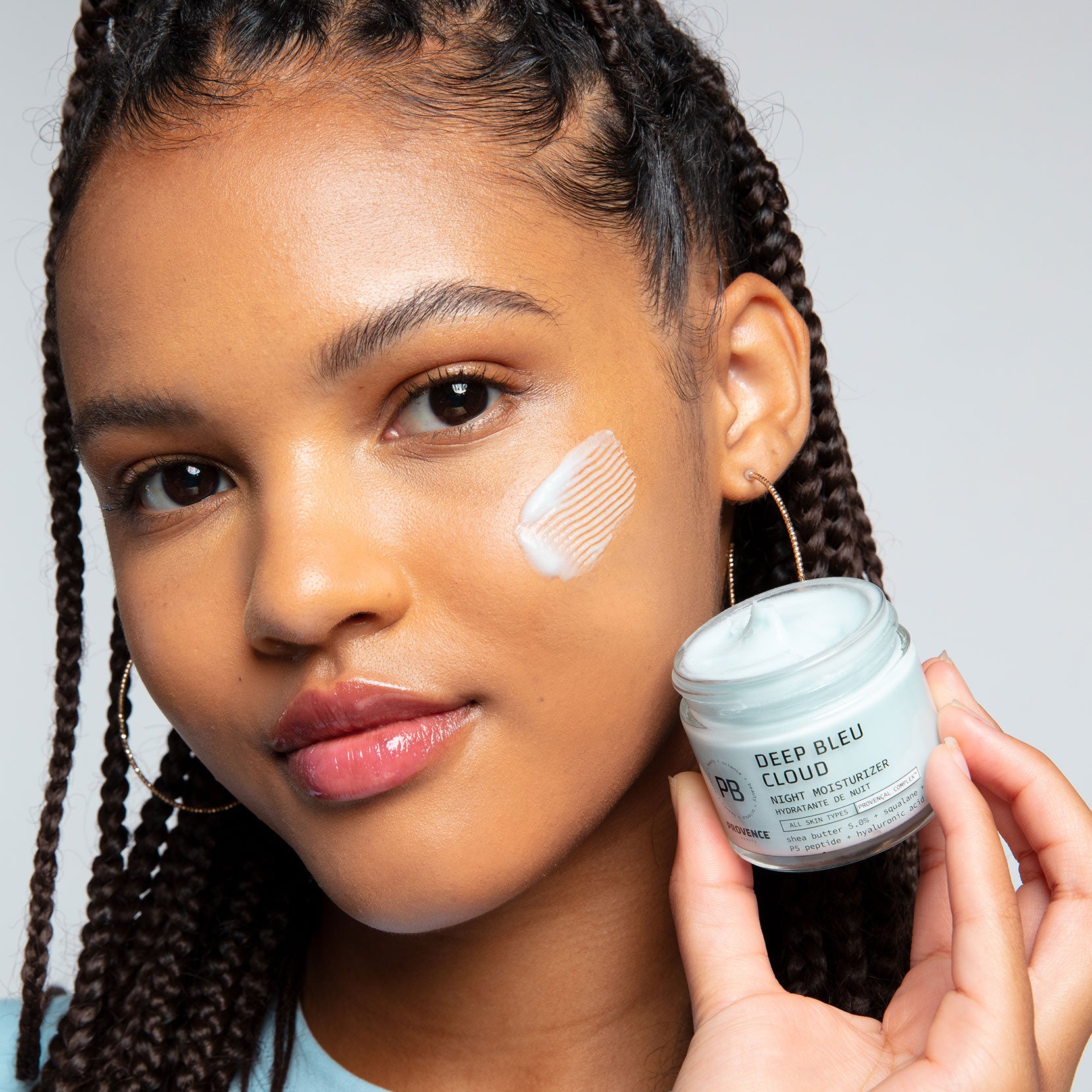 A model with glowing skin and braids posing with the Deep Bleu Cloud Night Moisturizer with a smoosh of the cream on her cheek. Due to its thickness there are streaks in the product from the spatula that smeared the product on her face.
