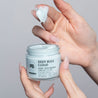 Image of a model's hands with long nails and freckles holding the Deep Bleu Cloud Night Moisturizer with two fingers that have been dipped in the cream showing the thick texture.