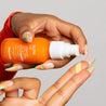 Image of a model's hands with long nails and freckles holding Glow Up Luminescent Facial Serum and pumping the product onto her fingers showing the liquid consistency.